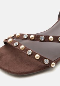 Καφέ σουέντ σανδάλι με δύο λουριά διακοσμημένα με εναλλασσόμενα χάντρες και διάφανα στρας πάνω σε απλό φόντο.