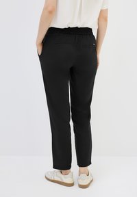 Svarta byxor med elastisk midja, med sidofickor och tapered ben. Materialet har en slät yta. Bärs med vita sneakers.