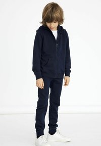 Hoodie zippé bleu marine avec poches avant, associé �à un pantalon bleu marine assorti et des baskets blanches. Tissu doux et extensible avec une texture lisse.