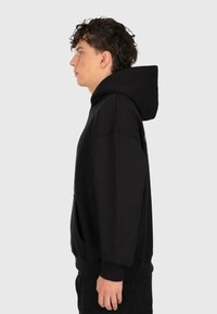 Young man in black oversized hoodie standing in profile against a plain light background.