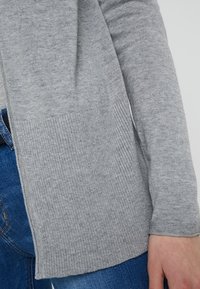 Ljusgrå kofta i mjukt stickat material, med ribbade detaljer och en avslappnad passform, kombinerad med blå denimjeans.