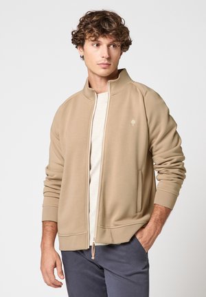 Jeune homme portant une veste beige zippée sur une chemise blanche, la main dans la poche, regardant sur le côté devant un fond uni.
