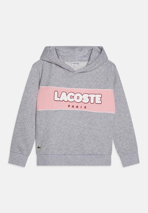 Le hoodie gris présente une bande horizontale rose avec "LACOSTE" et "PARIS" imprimés en lettres grasses. Matière douce, poche kangourou et capuche ajustable.