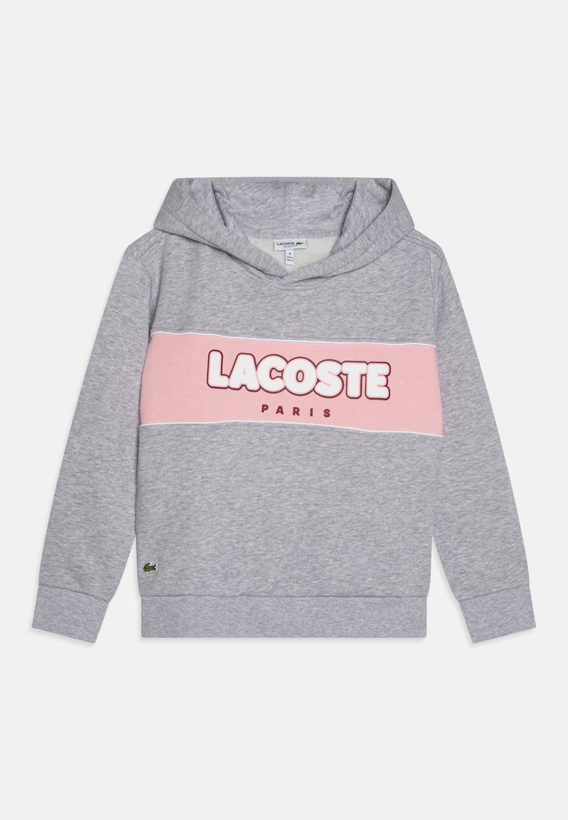 La sudadera gris presenta una franja horizontal rosa con "LACOSTE" y "PARIS" impresos en letras negritas. Material suave, bolsillo canguro y capucha ajustable.