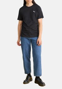 Schwarzes Baumwoll-T-Shirt mit kurzen Ärmeln und weißem Puma-Logo auf der Brust, kombiniert mit blauen Jeans und schwarzen Schnürschuhen.