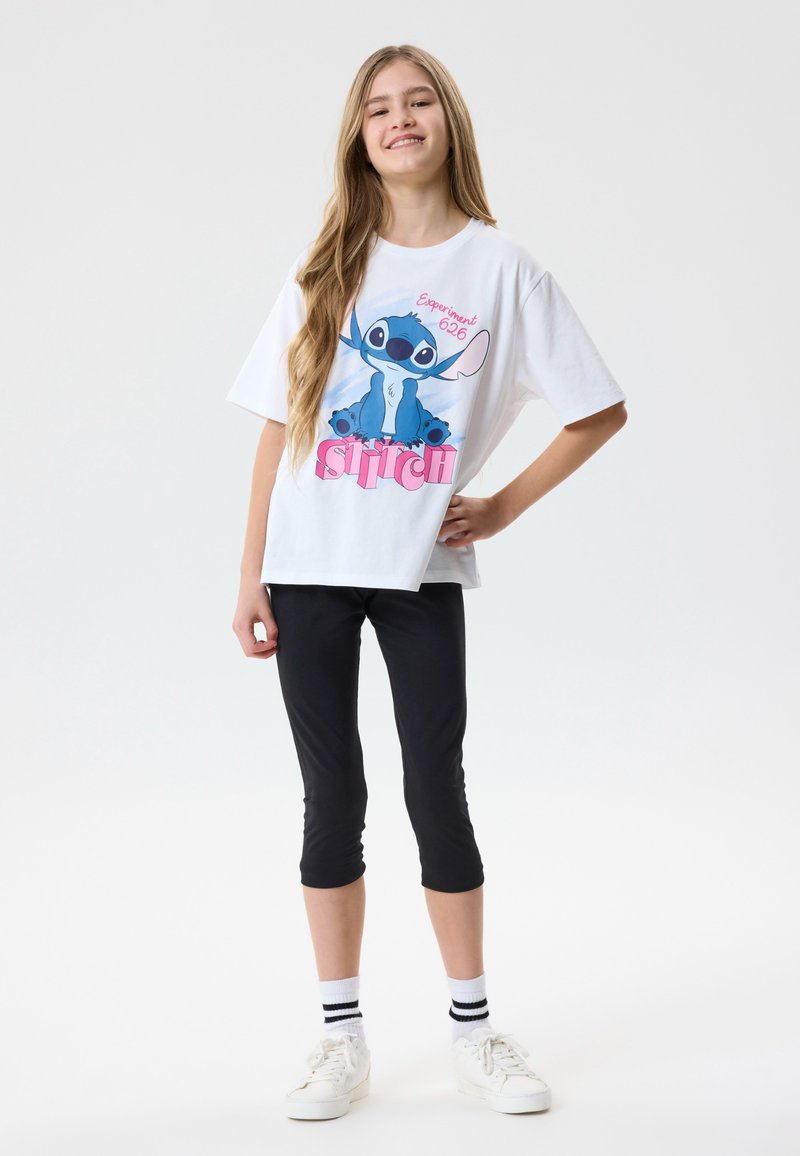 Jeune fille souriante, debout avec une main sur la hanche, portant un T-shirt blanc avec un motif Stitch, des leggings noirs courts, des baskets blanches et des chaussettes rayées.