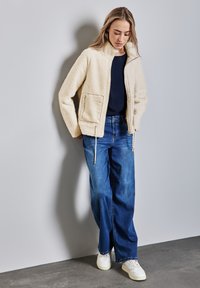 Veste en fausse fourrure de couleur crème avec un col haut, dotée d'une fermeture éclair à l'avant et de poches latérales, associée à un jean large bleu et des baskets blanches.