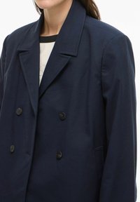 Femme portant un blazer bleu marine à double boutonnage avec des boutons noirs sur un haut clair à col noir.