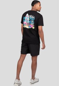 Camiseta negra con un diseño gráfico colorido en la parte posterior, que presenta elementos tropicales y texto. Combinada con pantalones cortos negros y zapatillas de deporte de color gris claro.