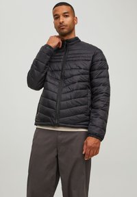 Svart, quilta puffjakke med høy krage og full glidelås, med elastiske mansjetter og en blank tekstur. Brukt over lyse klær.