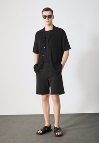 Chemise noire à manches courtes côtelée, t-shirt noir, shorts noirs et sandales noires avec des bretelles croisées. Modèle portant des lunettes de soleil noires.
