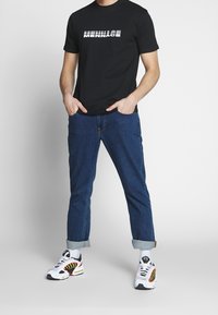 T-shirt noire avec le texte "MENNACE" en rayures blanches, associée à un jean bleu à revers et des baskets multicolores avec un design en maille.