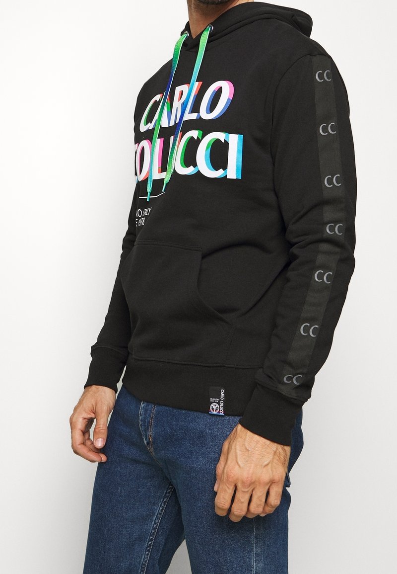 Homme portant un sweat à capuche noir avec le texte coloré « Carlo Colucci », des cordons verts et bleus et des logos « CC » sur la manche, associé à un jean bleu.