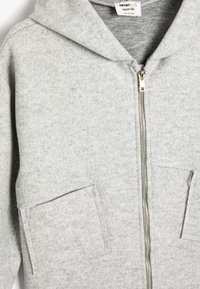 Sudadera gris con cremallera hecha de material de felpa suave. Presenta un gran bolsillo frontal y dos bolsillos laterales. Incluye una capucha con cordón.