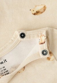 Jasnobeżowy bodziak z bawełny z drobnymi wzorami jeża, z zapięciami na napy i miękką fakturą. Etykieta pielęgnacyjna częściowo widoczna od wewnątrz.