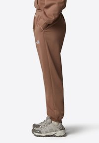 Bruna sweatpants i mjukt material, med elastiska mudd vid bensluten, sidofickor och en liten vit logotyp på vänster lår. Matchade med grå sportskor.