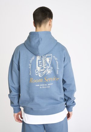 Person iført en blå hættetrøje med hvid og gul tekst og grafik af en vinflaske, glas, lys og mad, set bagfra.
