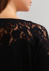 Pull noir avec des détails en dentelle florale dans le dos, des textures contrastées et un col rond. Tissu doux et léger.