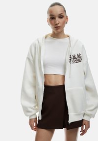 Ung kvinna klädd i en vit kort topp, en överdimensionerad vit zip-up hoodie med nallebjörnstryck och en mörkbrun minikjol, står mot en vit bakgrund.