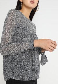 Blusa a maniche lunghe trasparenti in bianco e nero con motivo a righe, scollatura tonda e dettaglio con fiocco in vita. Design geometrico sottile su tutta la superficie.