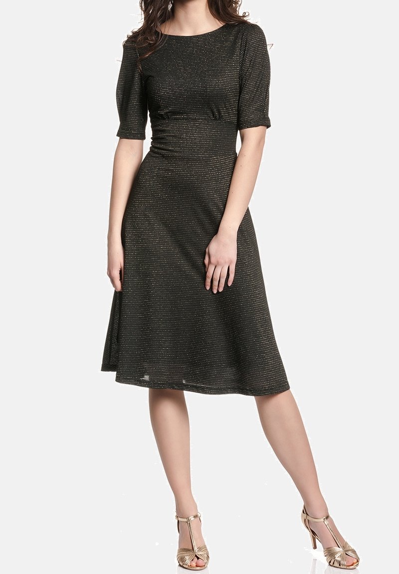 Femme portant une robe noire mi-longue avec un léger éclat et des manches mi-longues, associée à des sandales dorées à talons hauts ouvertes à l’avant.