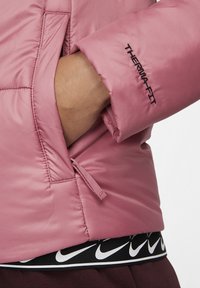 Rosa isolerad jacka med Thermafit-logotyp, med en närbild på en hand i en sidoficka. Mjuk tyg och sömnadsdetaljer synliga.