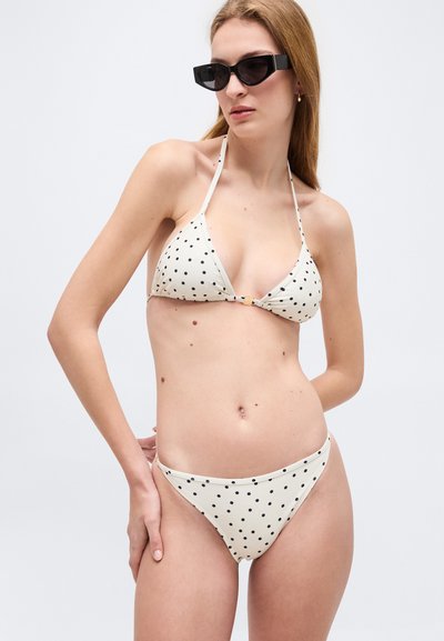 Γυναίκα που φοράει λευκό μπικίνι με μαύρες πουά και μαύρα γυαλιά ηλίου, στέκεται με το ένα χέρι στη μέση σε απλό φόντο.