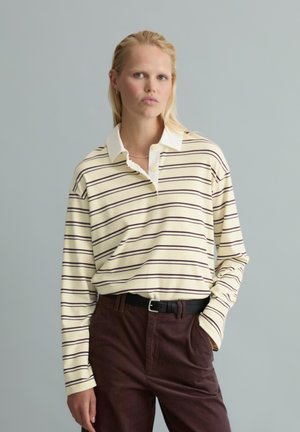 Blonde Person trägt ein beige-schwarz gestreiftes langärmeliges Polohemd, das in dunkelbraune hoch taillierte Hosen mit schwarzem Gürtel gesteckt ist.