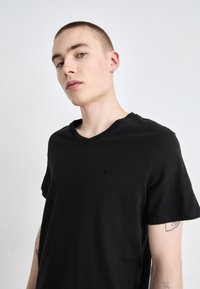 Fekete pamut póló V-nyakkal, rövid ujjakkal és egy kis logóval a mellkasán. Az anyag puhanak tűnik, sima felülettel.