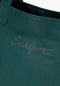 Bolsa de lona verde con un destacado logo "Scalpers" en letra cursiva morada. Tela texturizada con borde superior estructurado y asas reforzadas.