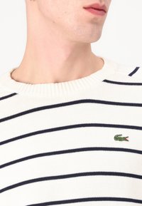 Sweater riscado em creme com linhas horizontais em azul-marinho, gola canelada e pequeno logótipo de crocodilo verde no lado esquerdo do peito. Tecido de malha texturizado.