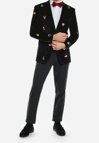 Man med svart sammetsblazer dekorerad med julmotiv, röd fluga, gråbyxor och svarta skor, med armarna i kors.