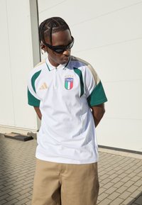 Biely polokošeľa s zelenými akcentmi, zipsový golier a erb Talianska. Zlaté pruhy na pleciach. Nosené s béžovými kraťasmi. Čierne slnečné okuliare.