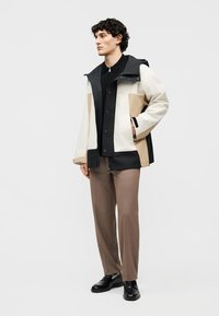 Άνδρας που στέκεται φορώντας μπουφάν με χρωματικά μπλοκ σε μπεζ, λευκό και μαύρο, μαύρο πουκάμισο, καφέ παντελόνι και μαύρα παπούτσια, κοιτάζοντας αριστερά.