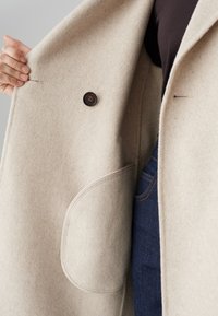 Hand houdt een beige wollen jas open en toont de binnenzak, knoop, donker shirt en de tailleband van spijkerbroek.