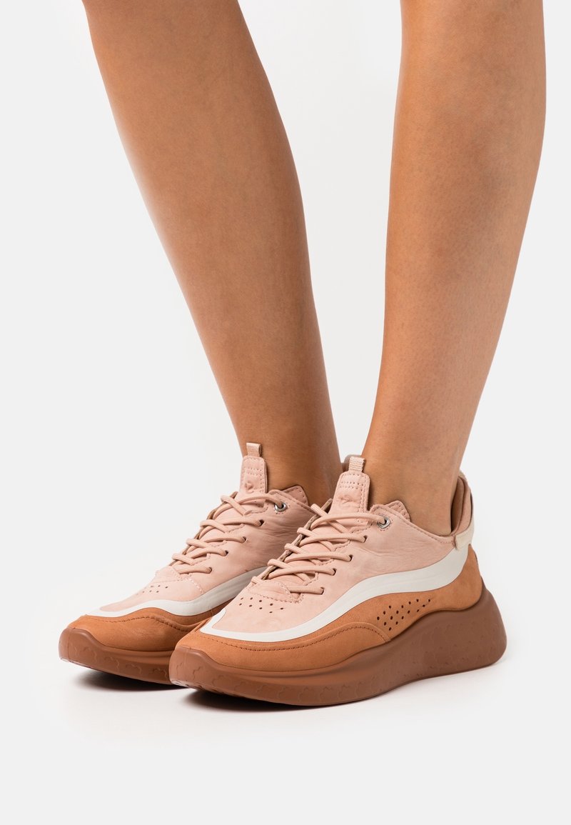Jambes portant des baskets lacées roses et marron avec des semelles épaisses sur un fond blanc uni.