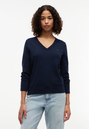 Femme portant un pull bleu marine à col en V avec des détails de boutons sur les manches et un jean bleu clair, sur un fond blanc uni.