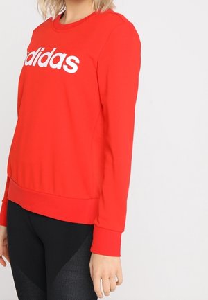 Kobieta w jaskrawoczerwonej bluzie Adidas z białym logo i czarnych, teksturowanych legginsach, stojąca na tle jasnego, jednolitego tła.