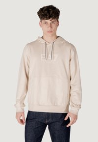 Felpa beige con cappuccio e cordino, tasca frontale e logo EA7 in rilievo. Realizzata in tessuto morbido con polsini a coste. Indossata con jeans scuri.