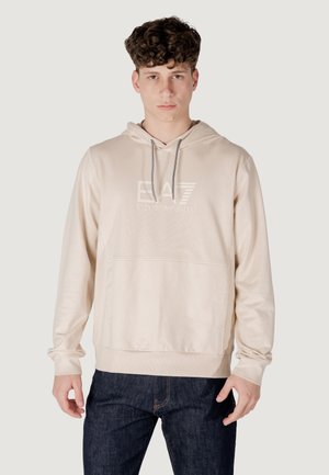 Felpa beige con cappuccio e cordino, tasca frontale e logo EA7 in rilievo. Realizzata in tessuto morbido con polsini a coste. Indossata con jeans scuri.