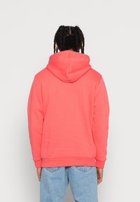 Korallrosa hoodie med en stor, justerbar huva, ribbade ärmslut och nederkant, tillverkad av mjukt tyg. Visad från baksidan.