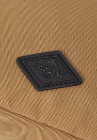 Borsa di tessuto color sabbia con superficie testurizzata, patch in pelle nera con il logo impresso "REPLAY 1981" e cuciture dettagliate.