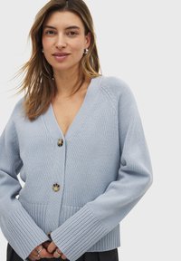 Cardigan en maille bleu clair avec un décolleté en V profond, des poignets côtelés et une fermeture à boutons sur le devant. Présente des accents texturés et une coupe décontractée.