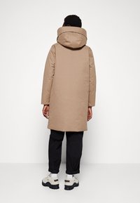 Tan parka med høj krave og hætte, der har en glat tekstur, lange ærmer og sidelommer, parret med sorte bukser og sneakers.