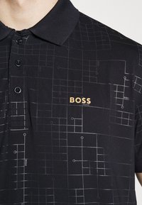 Black polo shirt with a geometric pattern in gray, featuring a three-button placket, textured collar, and a gold logo on the chest.