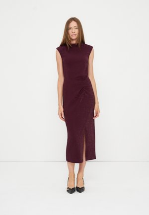 Woman with straight brown hair wearing a sleeveless dark purple midi dress with a front slit and black pointed-toe heels standing against a white background.