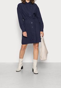 Abito bottonato blu navy con maniche lunghe, vestibilità slim e tessuto morbido. Ha un colletto ed è abbinato a stivali al ginocchio beige.