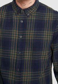 Mies, który ma na sobie button-up koszulę w kraciasty wzór w granatowo-zielono-brązowe kolory, z widocznym tatuażem na szyi nad kołnierzem.