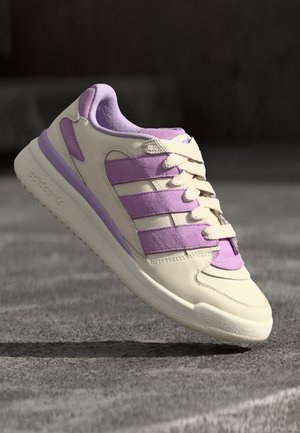 Kremowo-lawendowy but sportowy Adidas z zamszowymi trzema paskami, zawiązanymi sznurówkami i teksturowaną podeszwą, ustawiony pod kątem na rozmytym, ciemnym tle.