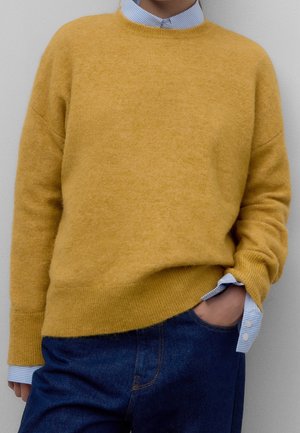 Maglione di lana giallo con orlo e polsini a coste, indossato sopra una camicia a righe blu chiaro, abbinato a jeans scuri.